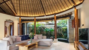 Amplia sala de estar con un sofá grande, una mesa de centro de madera y un televisor. La habitación cuenta con un techo alto con diseño tejido y ventanas grandes que ofrecen vistas de exuberante vegetación.