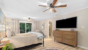 Une chambre à coucher comprenant un lit, un ventilateur de plafond, une commode, un téléviseur et une fenêtre avec des rideaux.