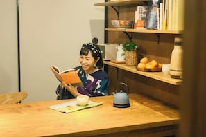 A person reading a book at a wooden table with a teapot, a cup, and a bowl of fruit.