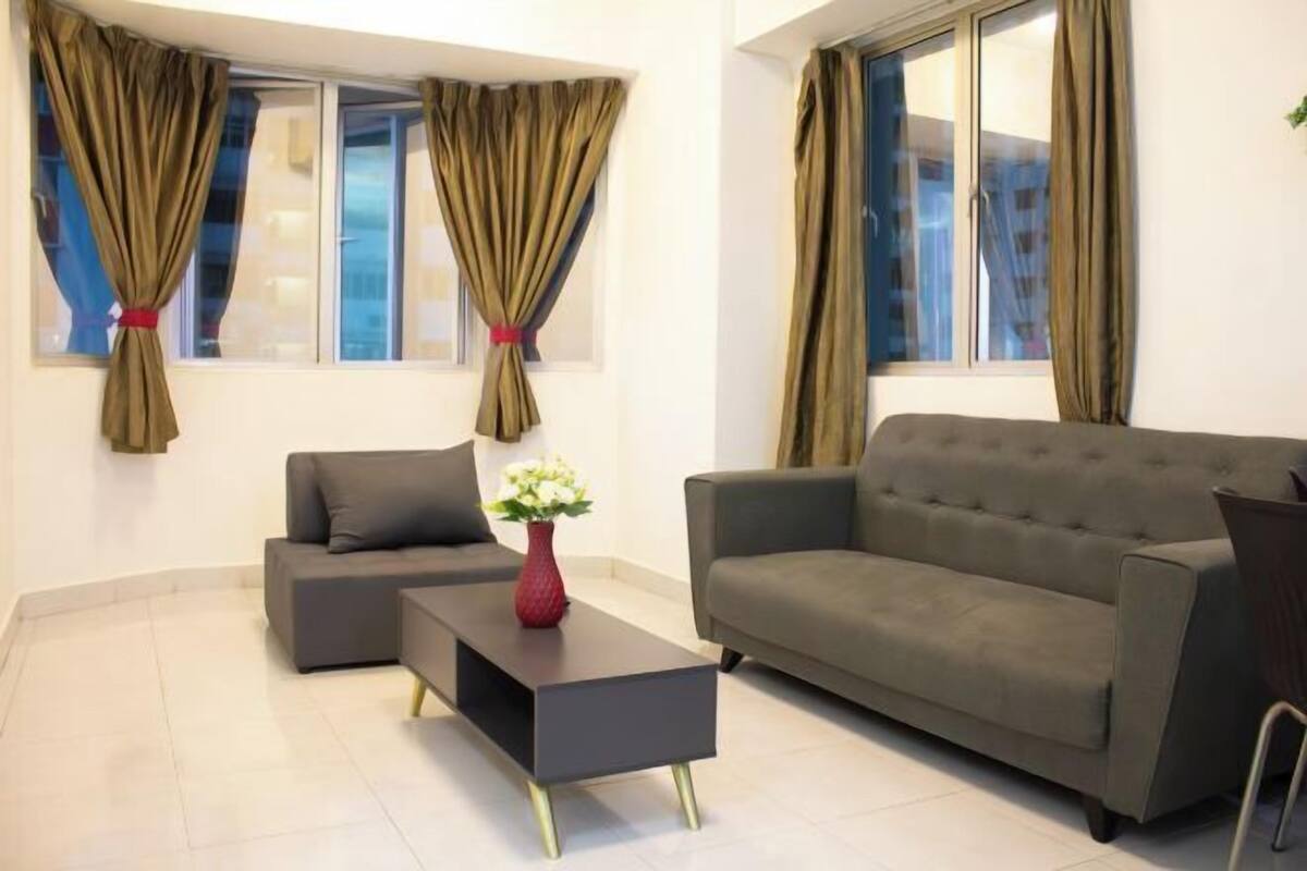 Ein modernes Wohnzimmer mit einem grauen Sofa, einem passenden Sessel und einem schwarzen Couchtisch. Zwei Fenster mit Vorhängen und eine Vase mit weißen Blumen auf dem Couchtisch.