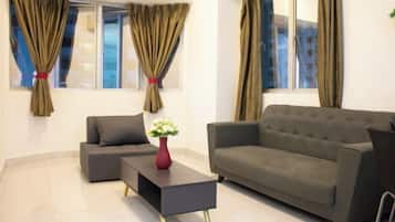 A modern living room with a grey sofa, a matching armchair, and a black coffee table. There are two windows with curtains, and a vase with white flowers on the coffee table.