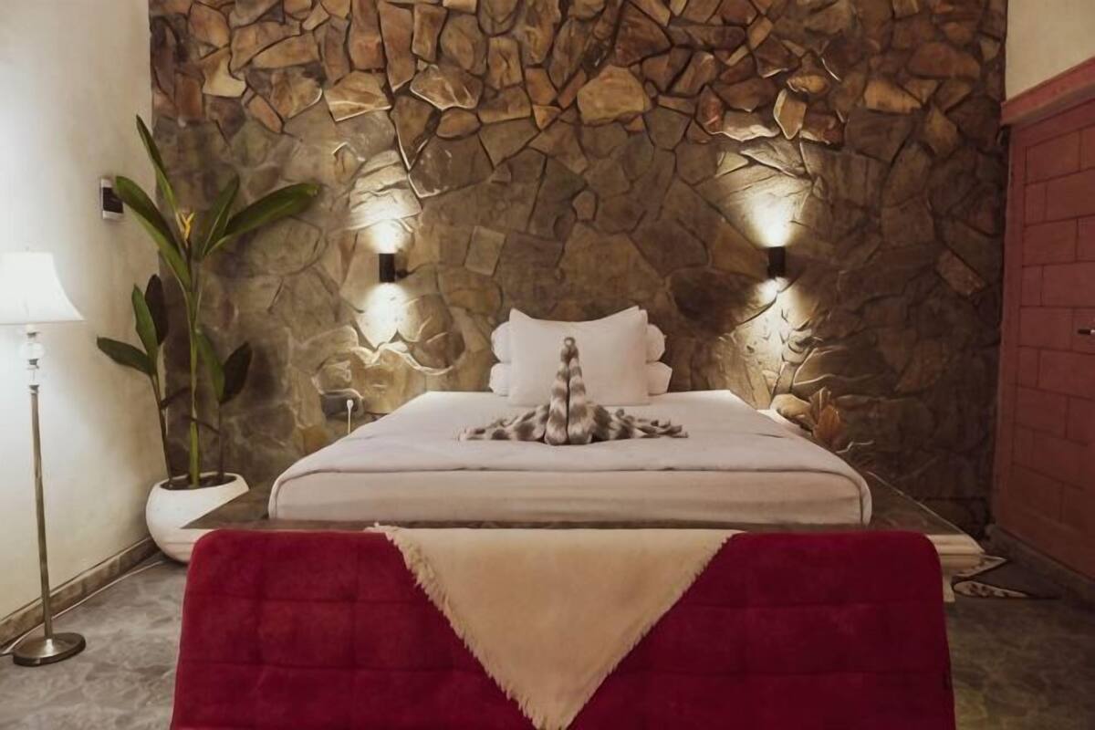 Quarto com parede de pedra, cama com almofadas e cabeceira estofada vermelha, e um abajur.
