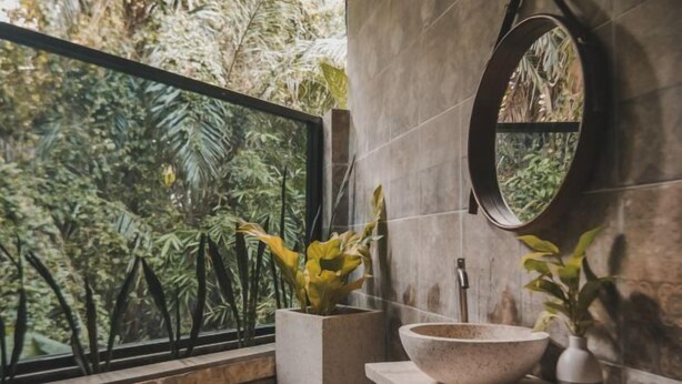 A bathroom with a large window, a round mirror, a stone sink, and a potted plant. - Villa Kamar Tamu Selomartani (Kalasan)
