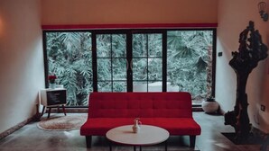 A red sofa, a white table, and a large window with a view of greenery. - Villa Kamar Tamu Selomartani (Kalasan)