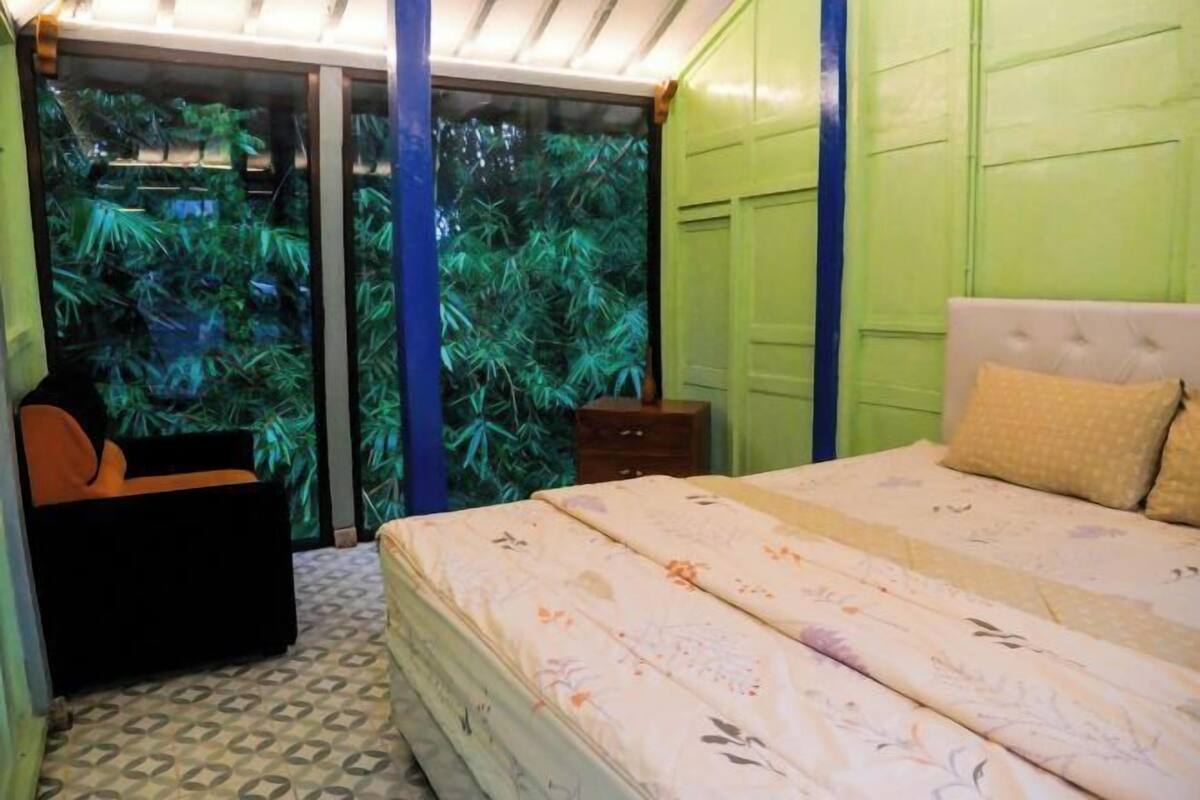 Quarto com cama, uma cadeira, uma mesinha e porta de vidro deslizante que dá para uma área verde externa.