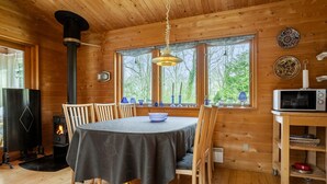 A cozy cabin interior with a dining table, chairs, a wood stove, a hanging light fixture, and a window with a view of trees. - 3 bedroom stunning home in Spøttrup (Spøttrup)