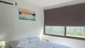 A neatly made bed with white linens and two towels, a bedside lamp, a painting of a sunset over water, and a window with a view of greenery.
