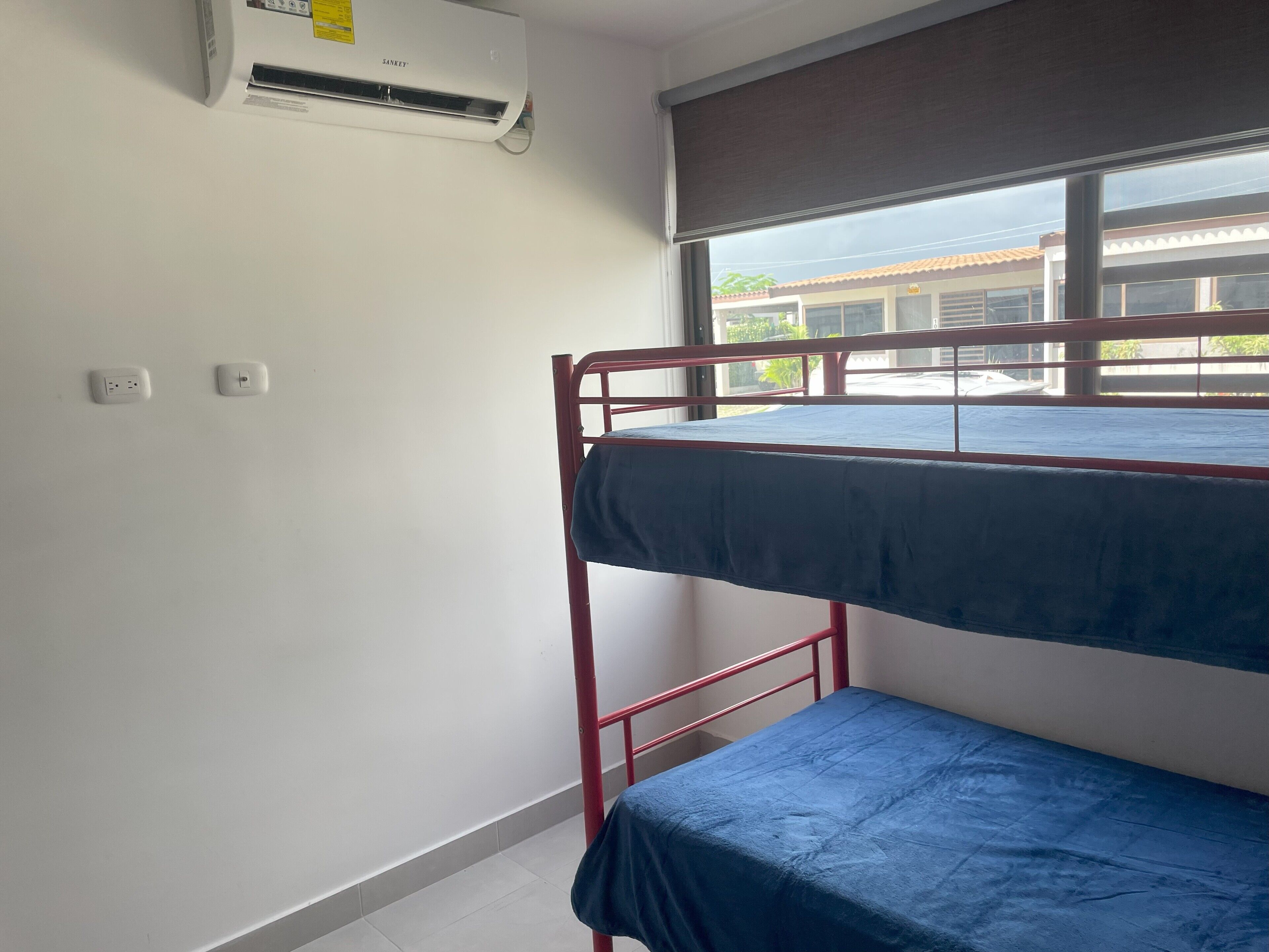Une chambre avec un lit superposé, une fenêtre, un climatiseur et deux prises électriques au mur.