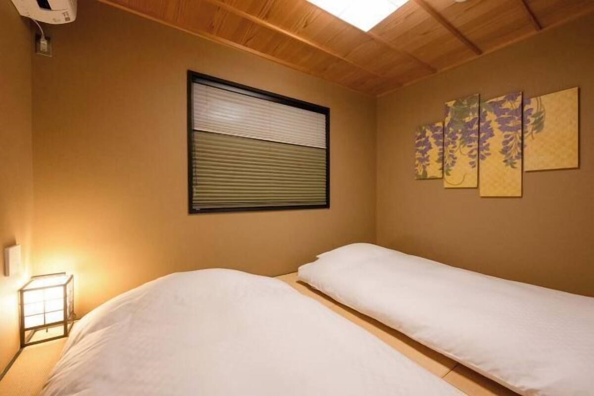 A room with two beds, a window with blinds, a wall-mounted light, and a set of paintings.