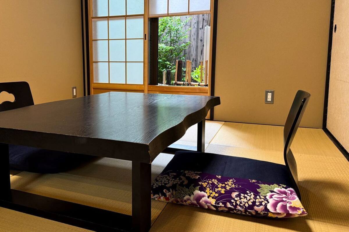 A tatami mat room with a low table and chairs, a shoji screen, and a view of greenery outside.