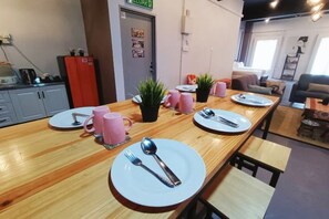 A long wooden table with plates, cutlery, and pink mugs in a modern room with a kitchenette and a bed in the background. - Melaka Riviera Studio 15 (Melaka Tengah)