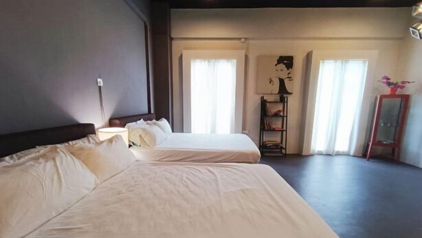 Quarto de hotel com duas camas, mesa de cabeceira, luminária, estante e janela com cortinas.