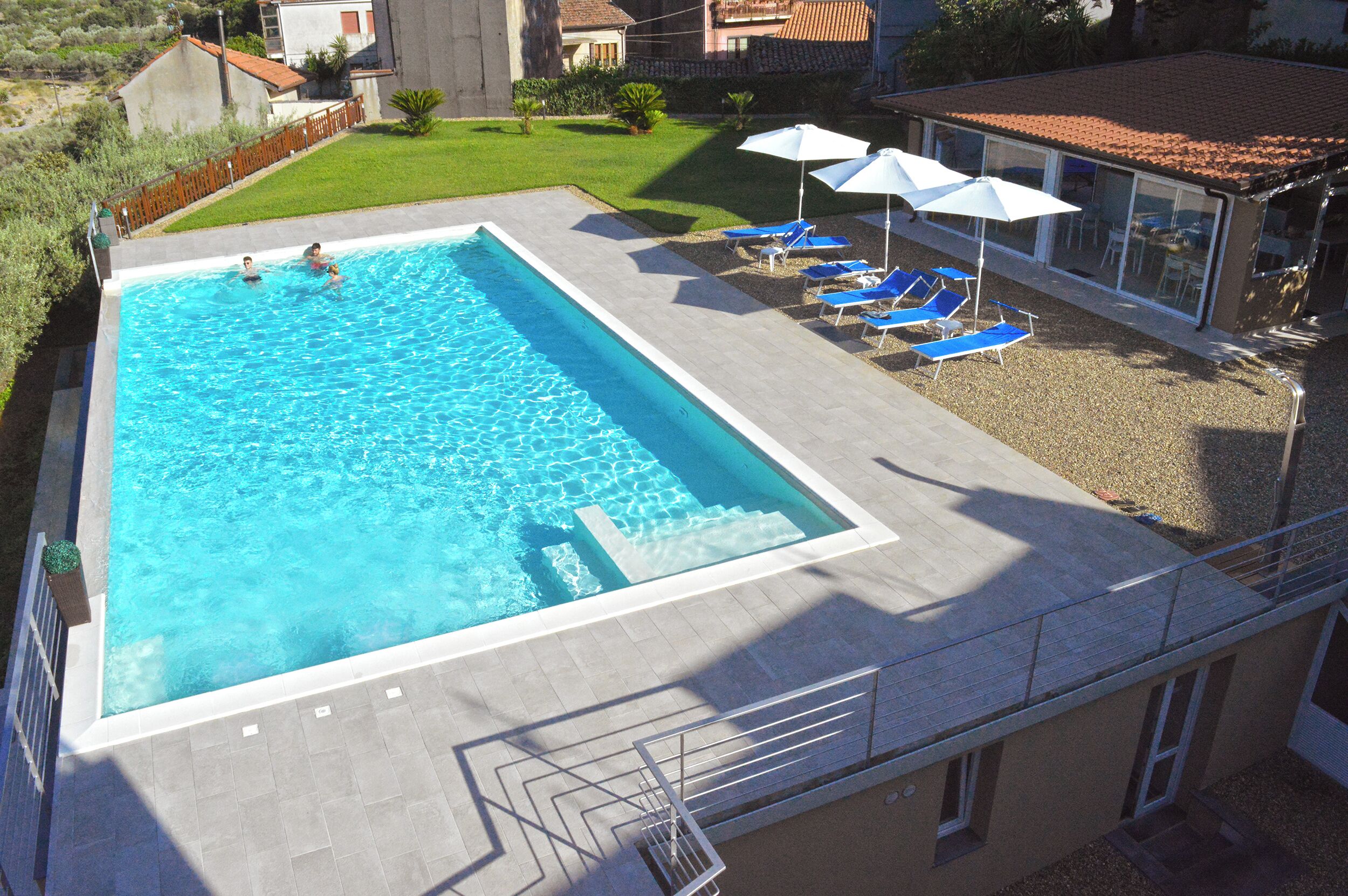 Una piscina rettangolare con bordo piastrellato, circondata da una piccola area erbosa e un edificio con tetto in tegole.