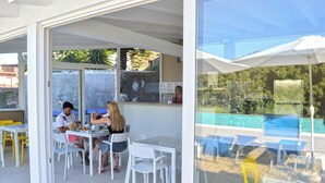 An outdoor dining area with white tables and chairs, a glass partition, and a view of a pool. - B&B Mauro Home bed&relax (Linguaglossa)