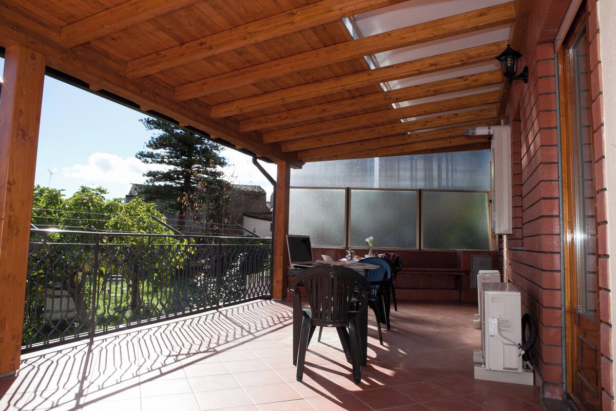 A covered outdoor area with a wooden ceiling, a table with chairs, and a refrigerator.