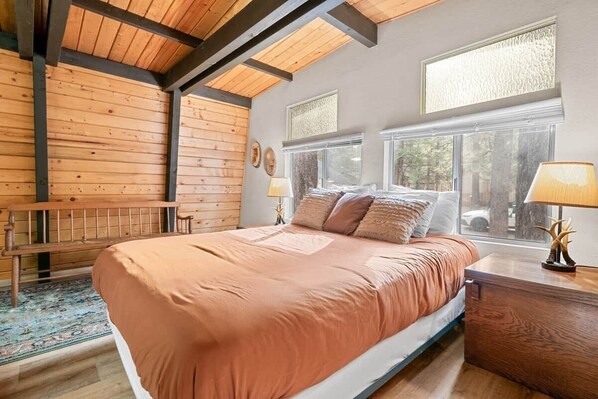 Un dormitorio con cama, mesitas de noche, un banco y un ventanal grande.