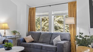 Ein modernes Wohnzimmer mit einem grauen Sofa, einem gelben Sessel und einem runden Couchtisch. Eine Lampe steht auf einem Beistelltisch, und ein Fenster mit Vorhängen ist zu sehen.
