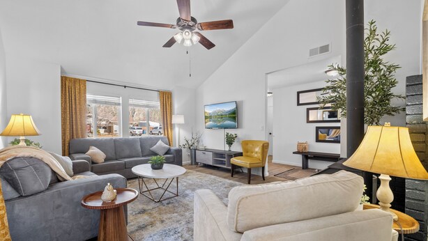 Ein modernes Wohnzimmer mit einem grauen Sofa, einem gelben Sessel und einem weißen Sessel. Es gibt einen Deckenventilator, einen an der Wand montierten Fernseher und einen Kamin.