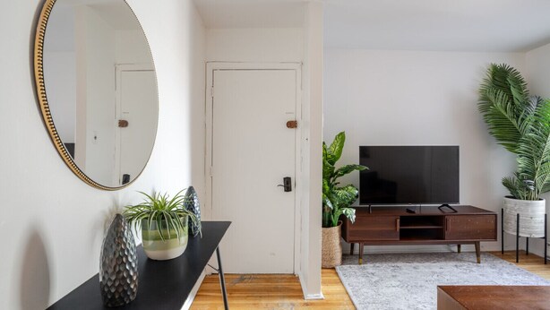A living room with a TV, wooden flooring, a black table with plants, a mirror, and a white door. - Modern 2BR in Plateau – Steps to Downtown (Montréal)