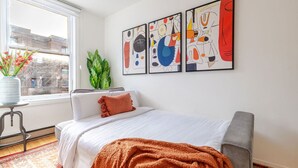 Een slaapkamer met een bed, een raam, een plant en abstracte kunst aan de muur.