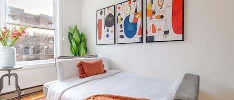 A bedroom with a bed, a window, a plant, and abstract art on the wall.