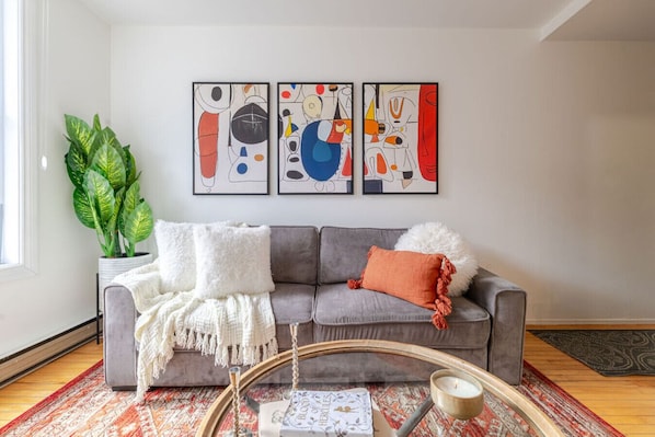 Un salon avec un canapé gris, des coussins blancs et orange, une plante en pot, des œuvres d’art abstraites accrochées au mur et une petite table sur laquelle est posée une bougie.