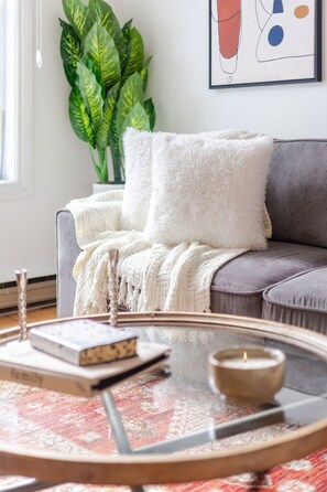 Una sala de estar acogedora con una mesita de centro redonda de vidrio, un sofá gris, un plaid blanco y esponjoso, y una planta verde grande.