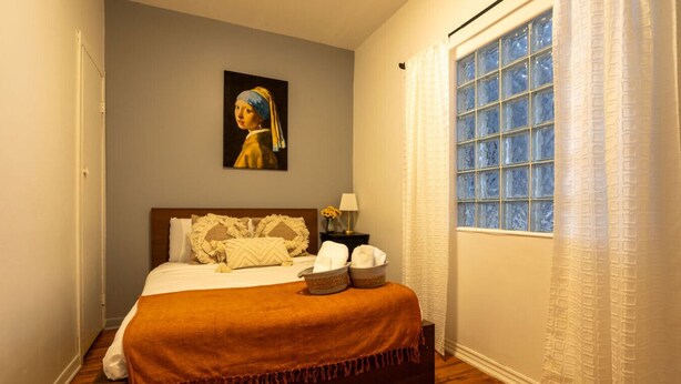 Une chambre à coucher bien rangée, avec un lit, des oreillers, une table de chevet avec une lampe et une fenêtre avec des rideaux.
