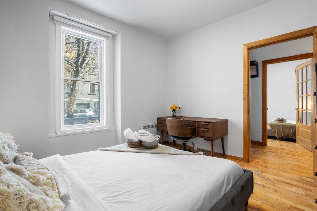 A bedroom with a bed, a desk, a chair, a window, and a door leading to another room.