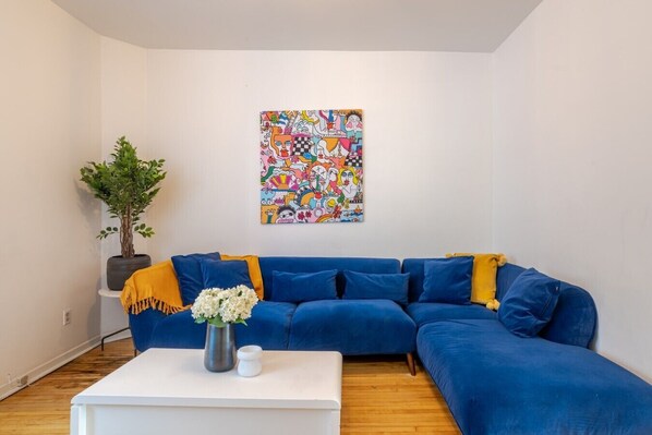 Una sala de estar con un sofá azul, una mesa de centro blanca, una planta en maceta y un cuadro abstracto colorido en la pared.