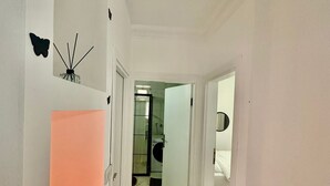 Un estrecho pasillo con paredes blancas, suelo de mármol y una puerta que da a un baño con cabina de ducha de vidrio.