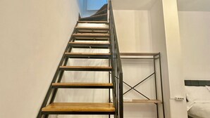 Una escalera estrecha con escalones de madera y barandillas metálicas que conduce a un dormitorio con cama y mesita de noche.