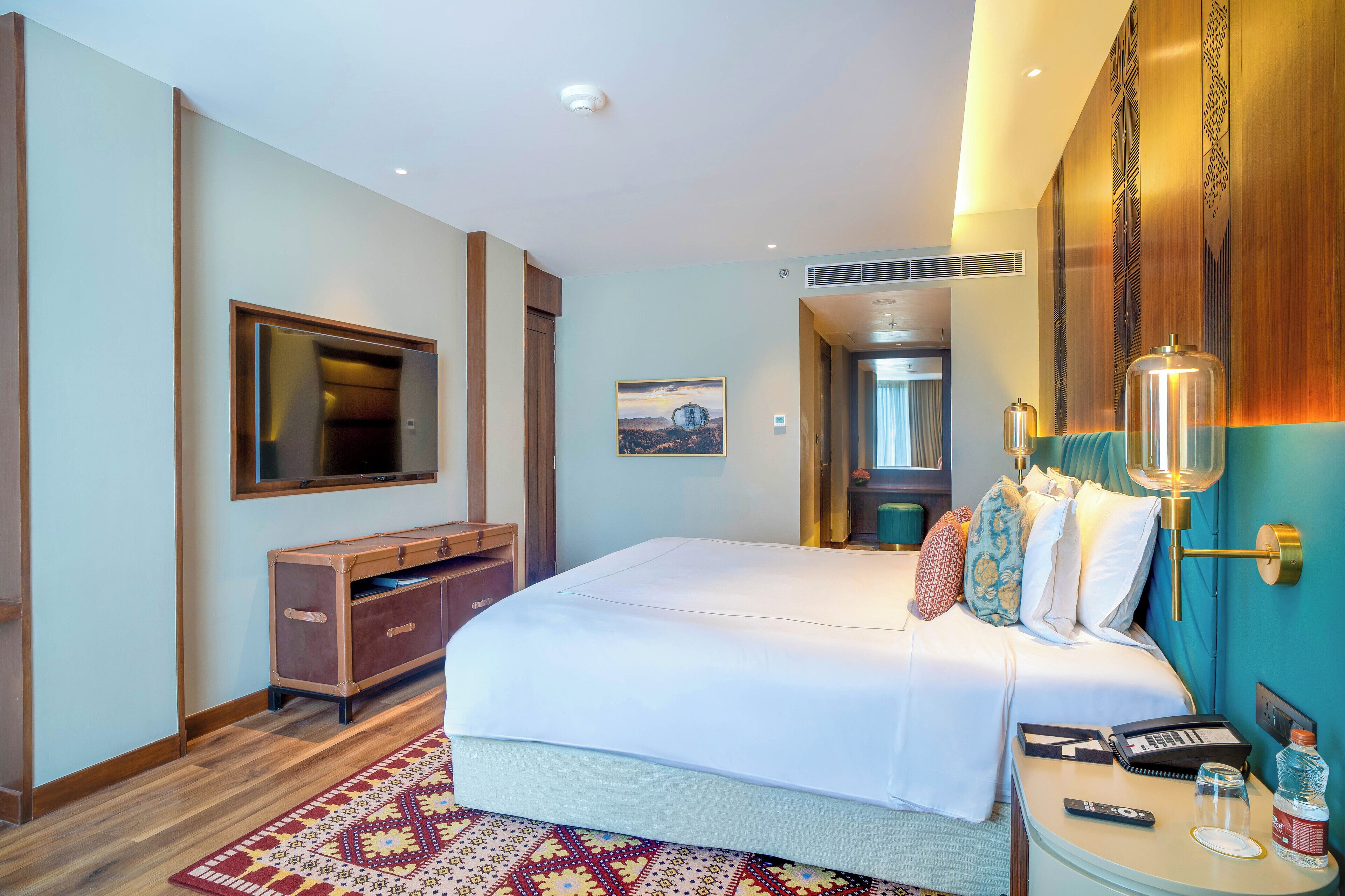A hotel room with a large bed, a TV, a desk with a phone and a lamp, and a wooden cabinet.
