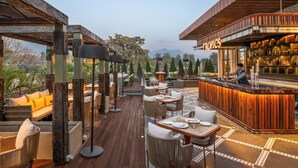 Un área de comedor al aire libre con mesas y sillas de madera, una barra con mostrador de madera, y una vista de árboles y montañas al fondo.