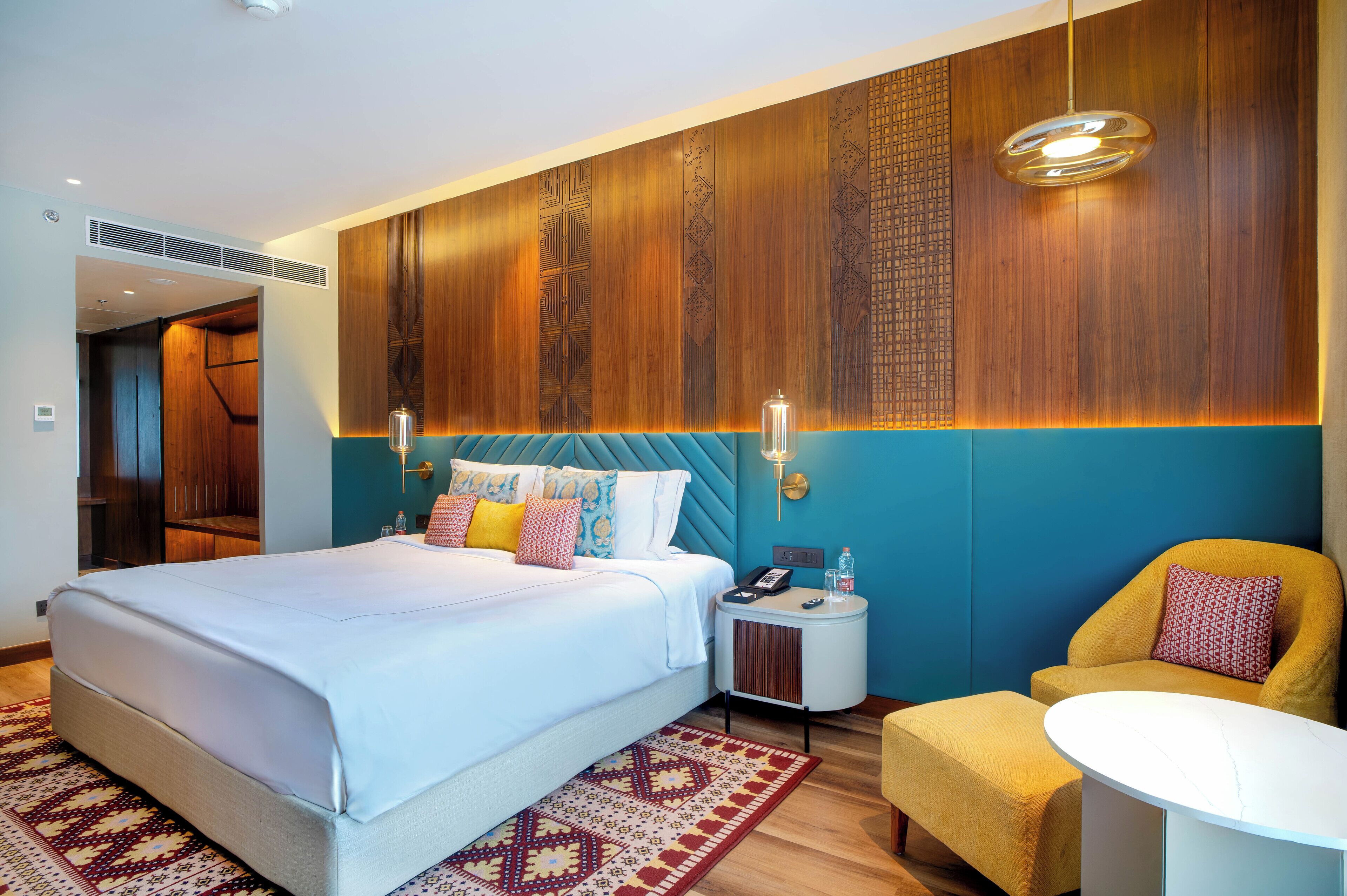 A modern hotel room with a large bed, a small side table, a yellow armchair, and a round table.