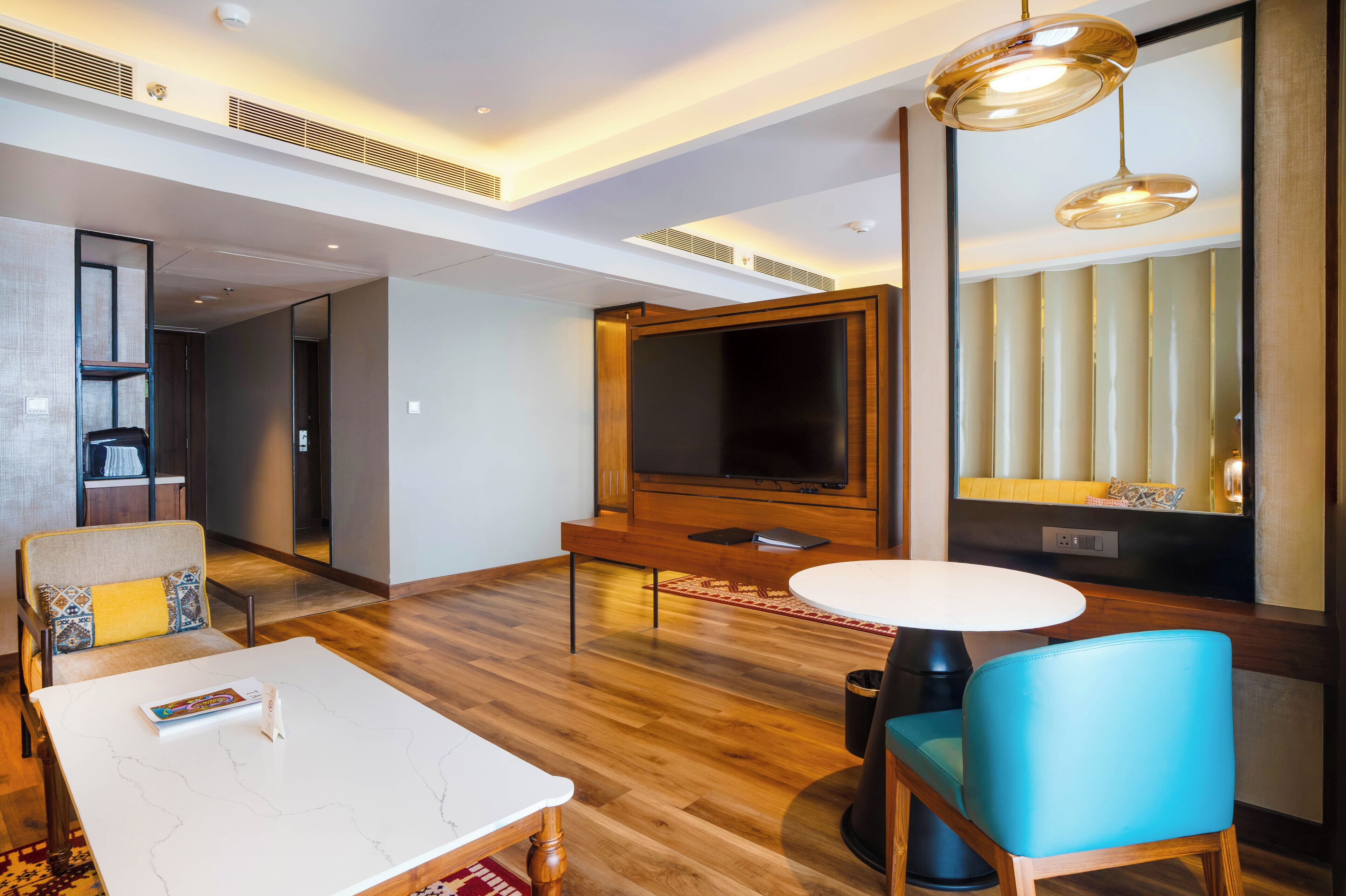 A modern hotel room with a wooden floor, a white marble table, a blue chair, a TV, and a large mirror.