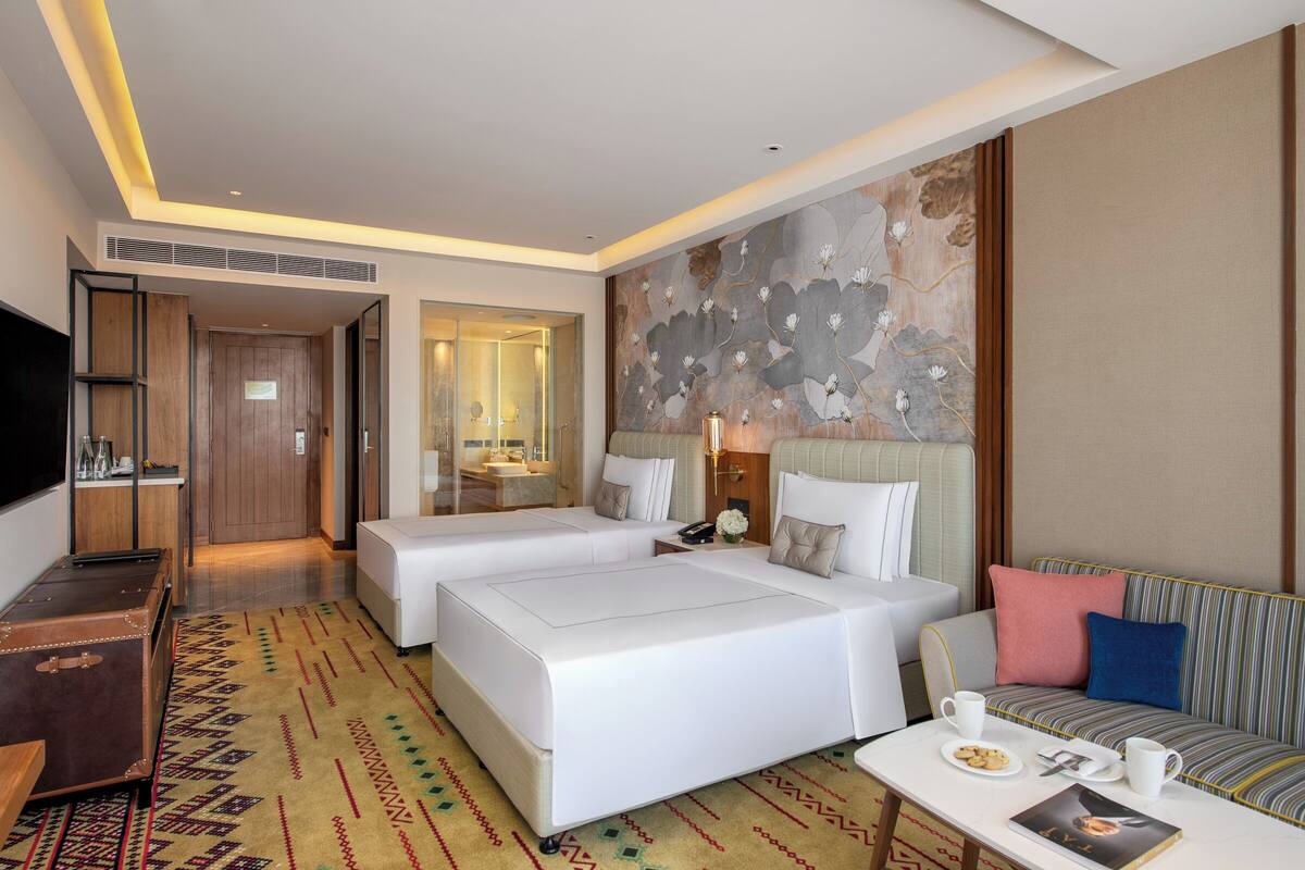 Habitación de hotel con dos camas, un sofá, una mesita con bandeja y un mural grande en la pared.