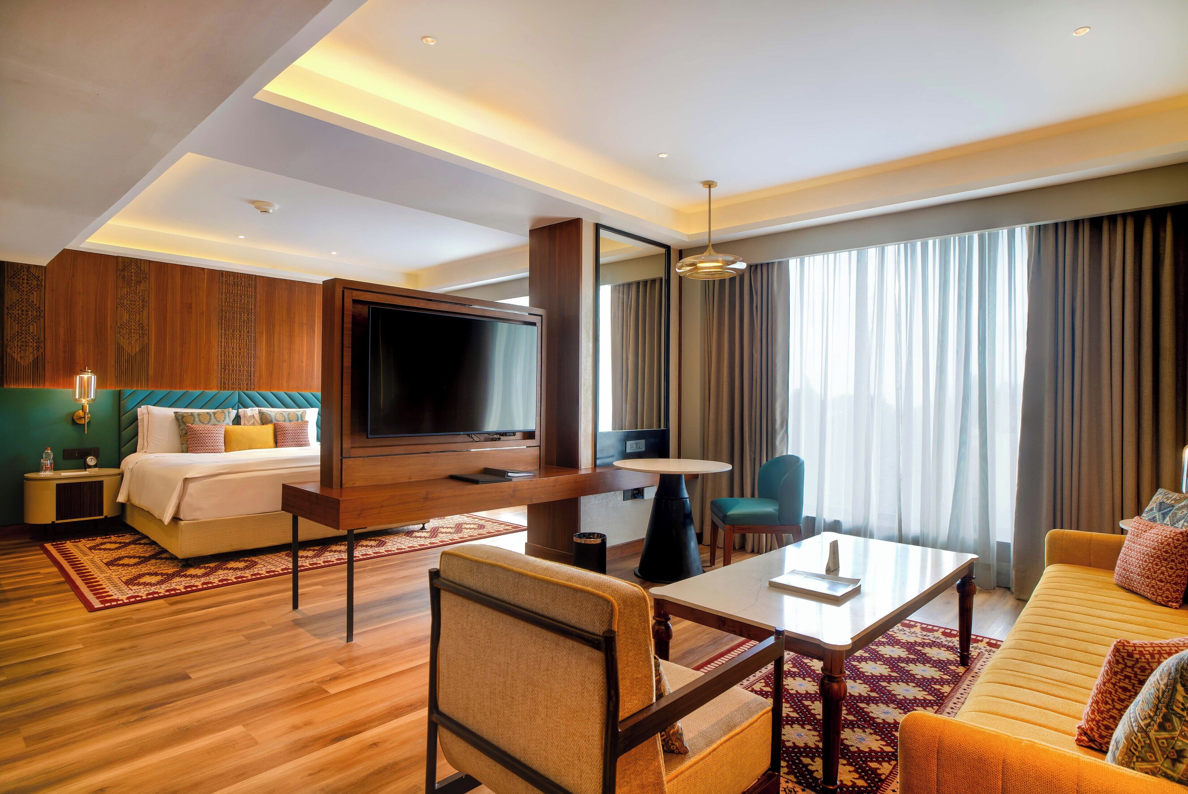 A hotel room with a bed, a TV, a dining table, a sofa, and a large window with curtains.