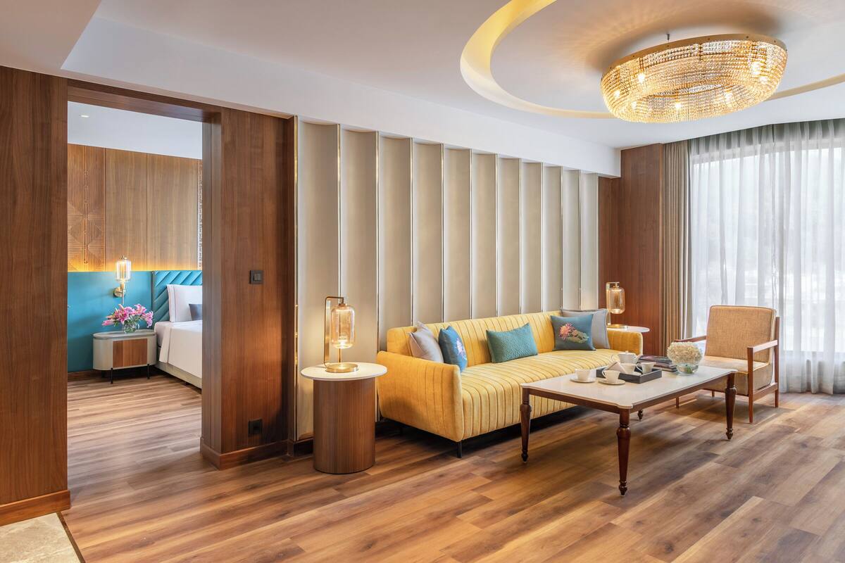Habitación de hotel moderna con suelo de madera, sofá con cojines, una mesa redonda, una silla, una lámpara y un amplio ventanal con cortinas.