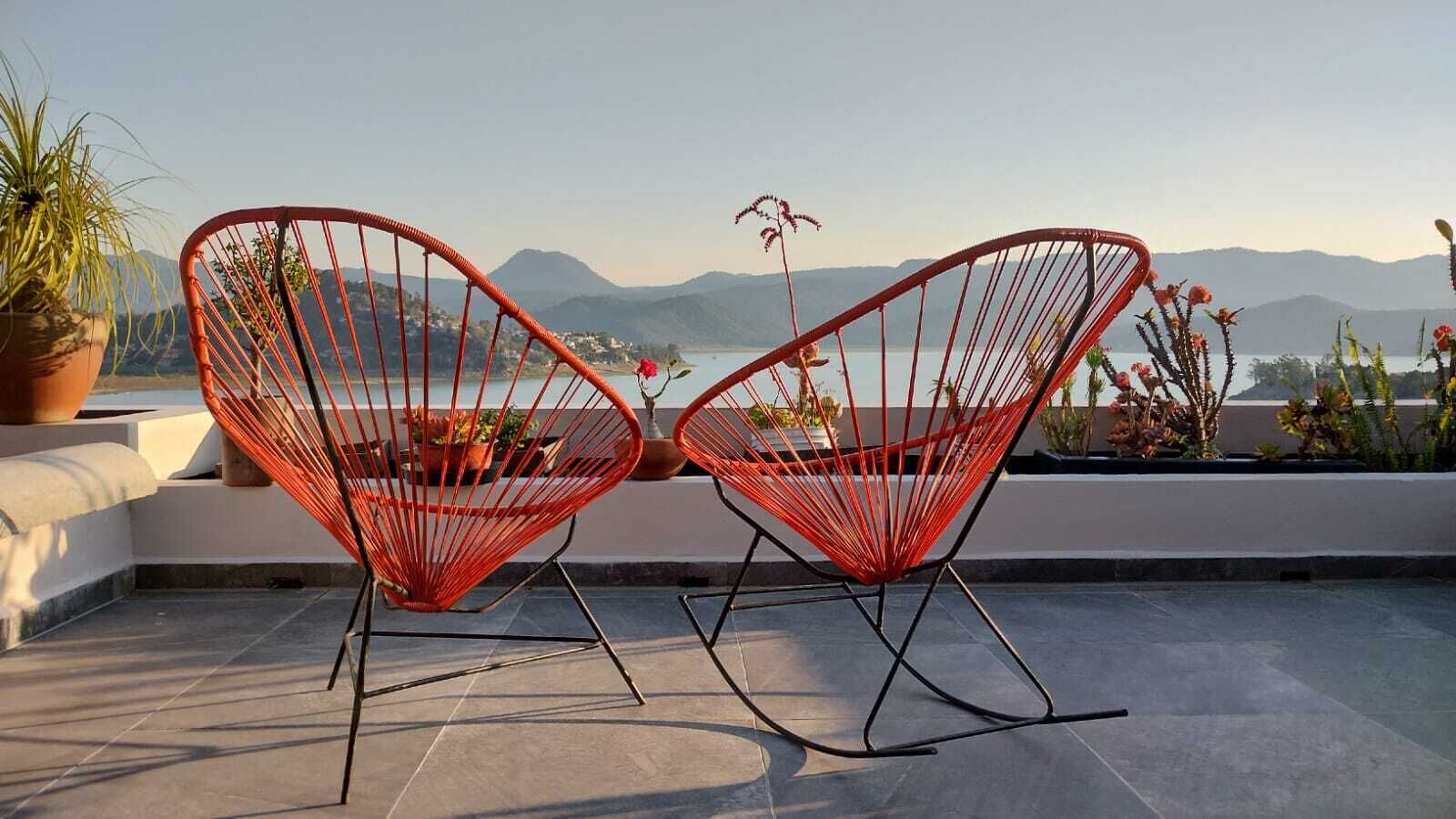 To røde stole med wire-stel på en tagterrasse med udsigt til bjerge og vand.