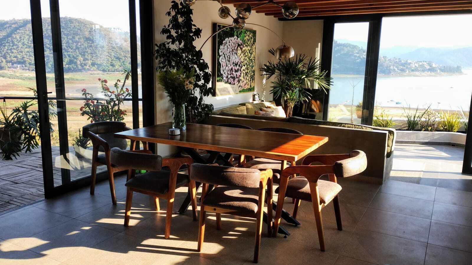 Uma zona de refeições com mesa e cadeiras de madeira, cercada por grandes janelas que oferecem vista para um lago e colinas.