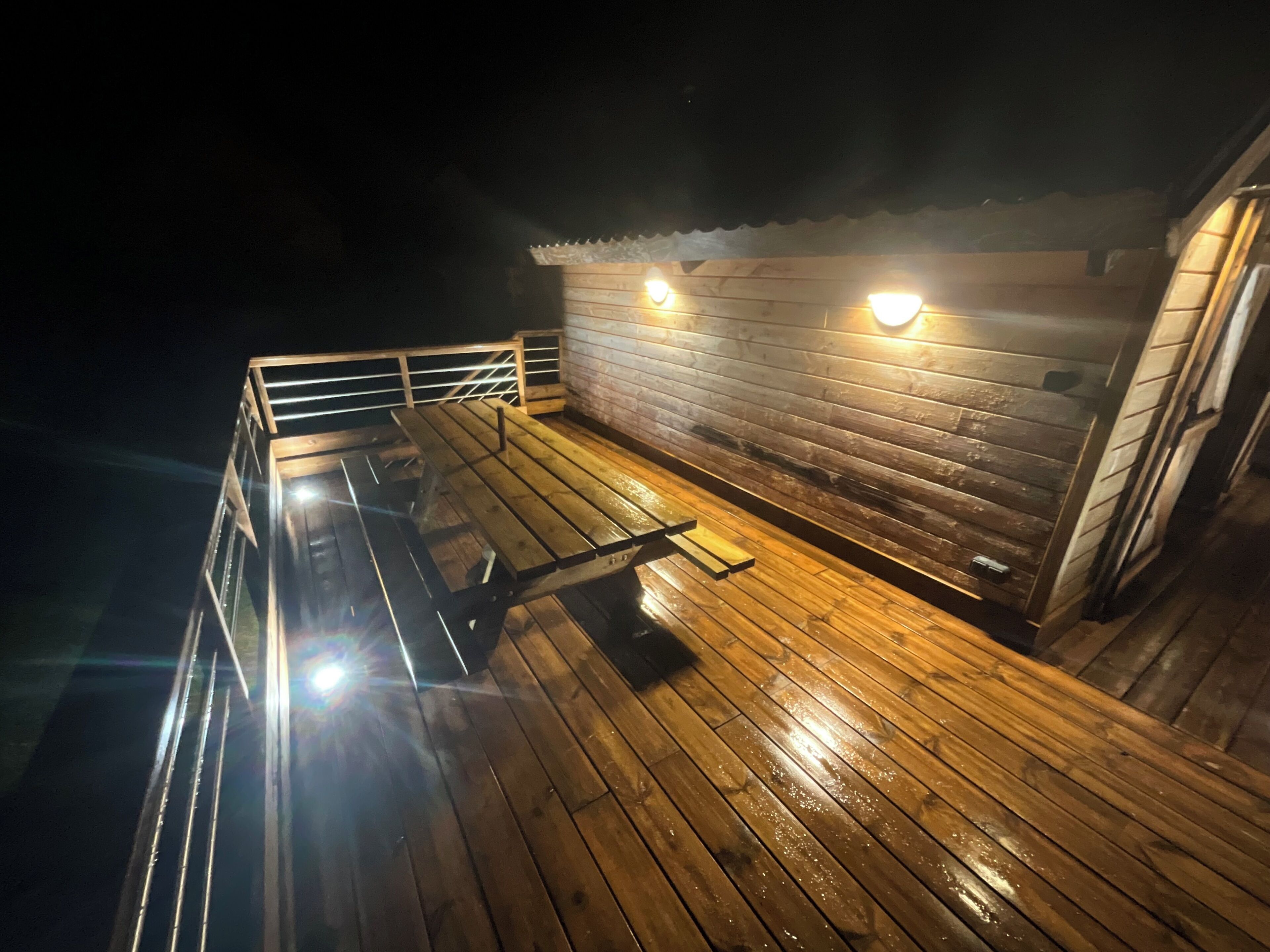 Una terraza de madera con una mesa de picnic y dos luces fijadas a la pared.