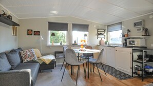 Un espace de vie compact comprenant un canapé, une table à manger, des chaises, un coin cuisine et une fenêtre donnant sur un espace verdoyant.