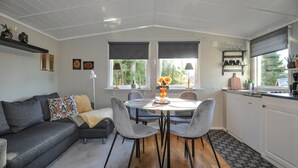 Un salon chaleureux avec un canapé gris, une table à manger avec quatre chaises et un coin cuisine doté d’une fenêtre et d’étagères.