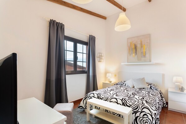 Un dormitorio con cama, mesitas de noche, un escritorio, una silla, una ventana con cortinas y un cuadro en la pared.