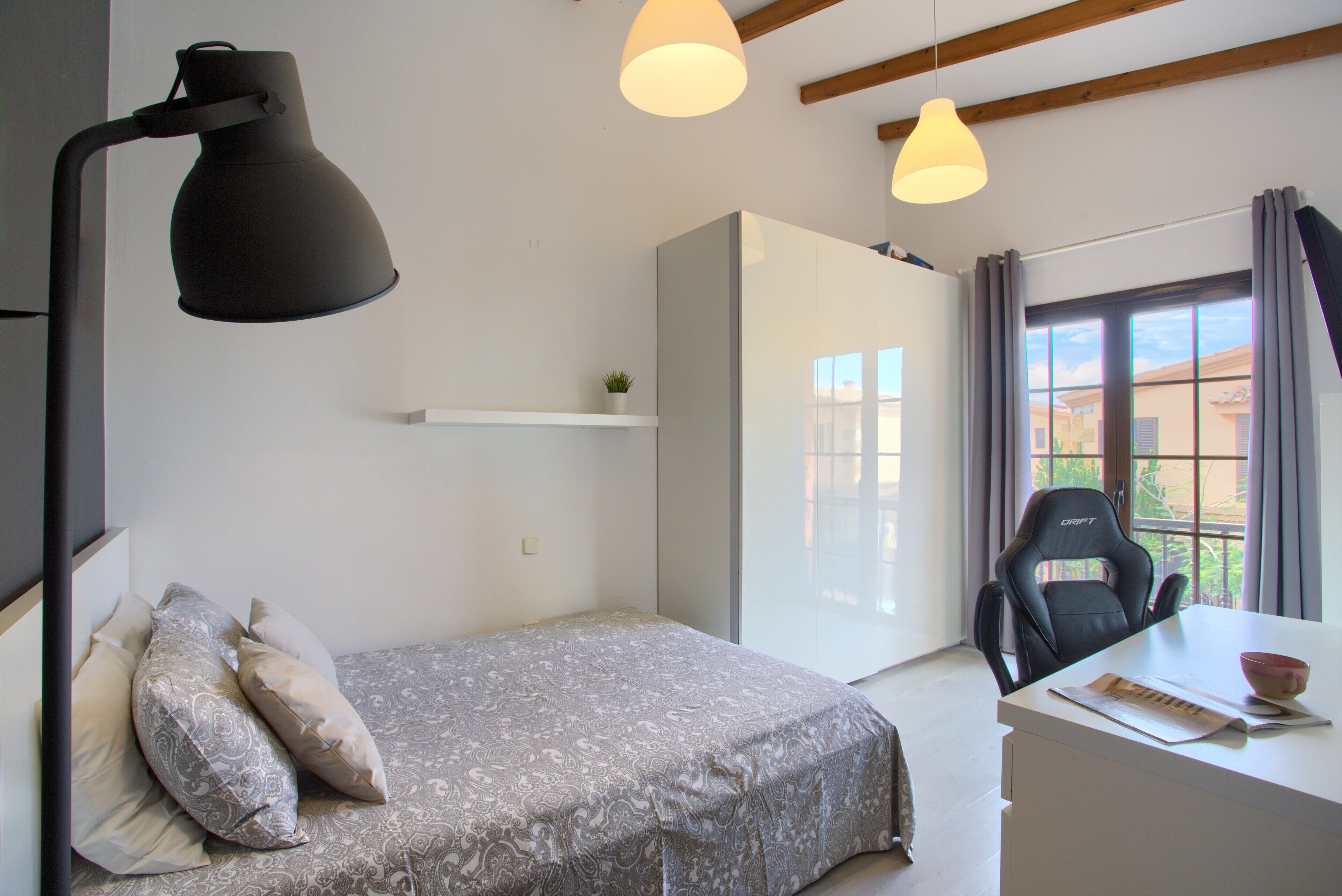 Une chambre à coucher comprenant un lit, une lampe de chevet, un bureau, une chaise et une fenêtre avec des rideaux.