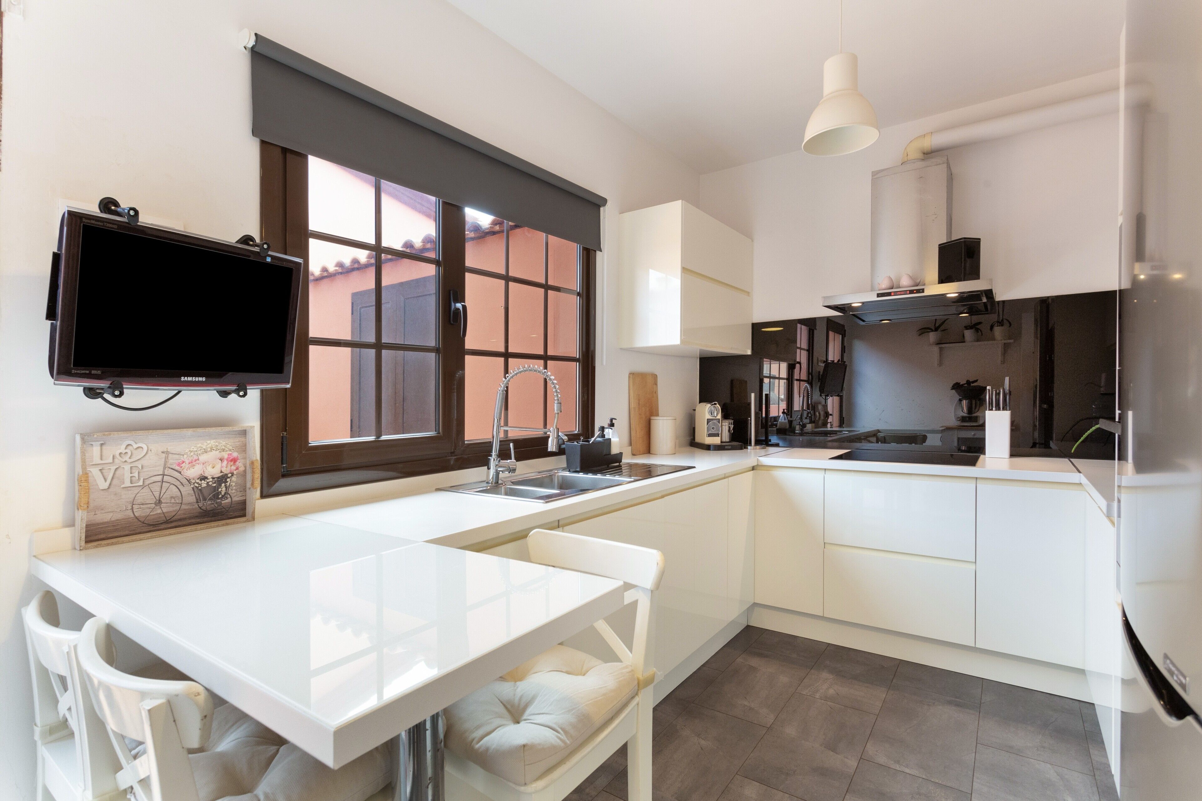 Eine moderne Küche mit integriertem Fernseher, weißen Schränken, einem großen Fenster und einem Essbereich mit weißem Tisch und Stühlen.