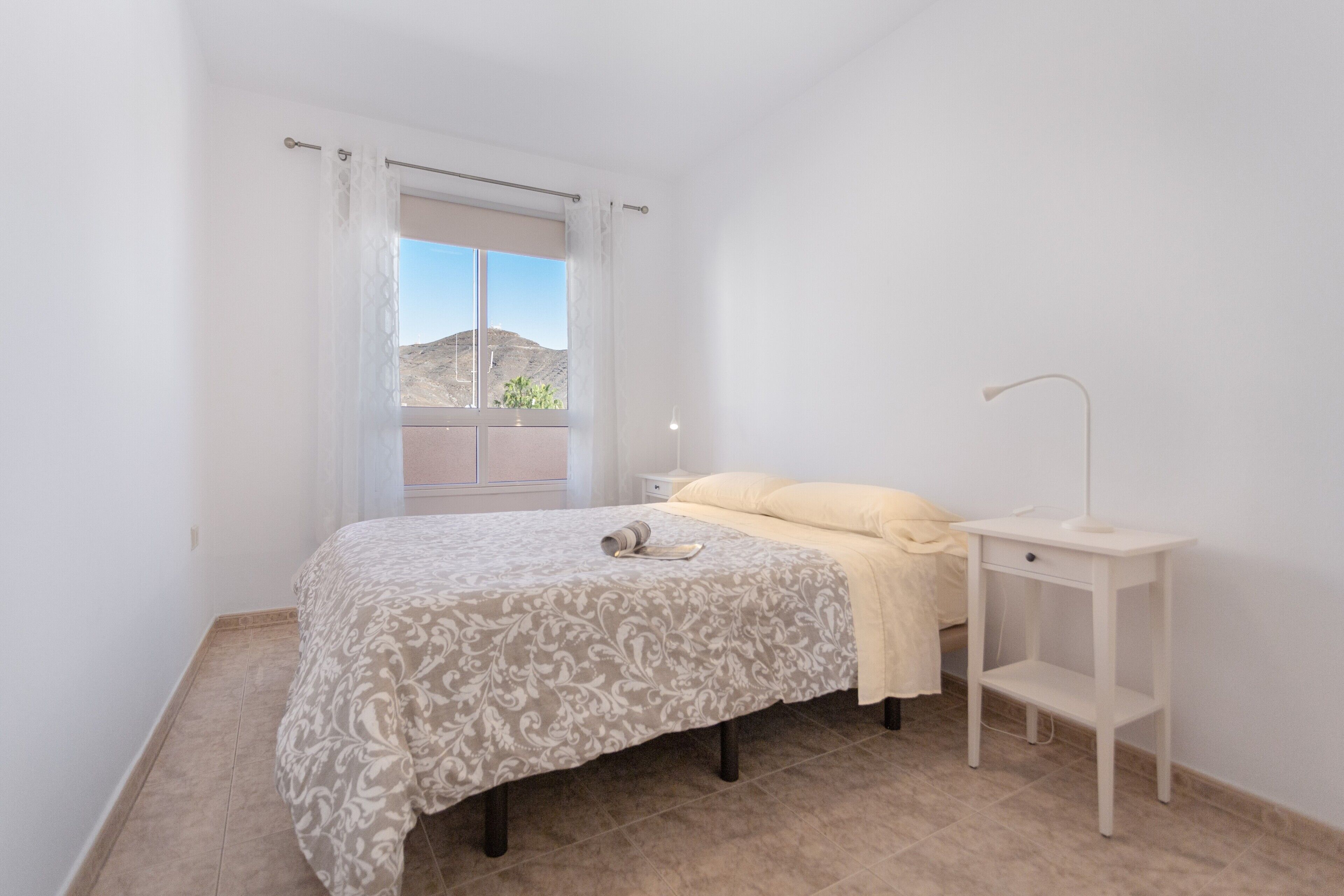 Un dormitorio con cama, mesita de noche, lámpara y ventana con vistas al exterior.