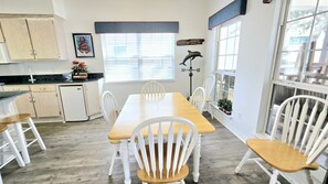 A dining area with a wooden table and white chairs, a kitchen with light wood cabinets, and a window with blinds. - Bear Point Bay Front Beach House! Sleeps 12 (Orange Beach)