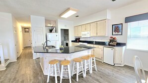 A modern kitchen with a central island, white bar stools, and wooden cabinets. - Bear Point Bay Front Beach House! Sleeps 12 (Orange Beach)